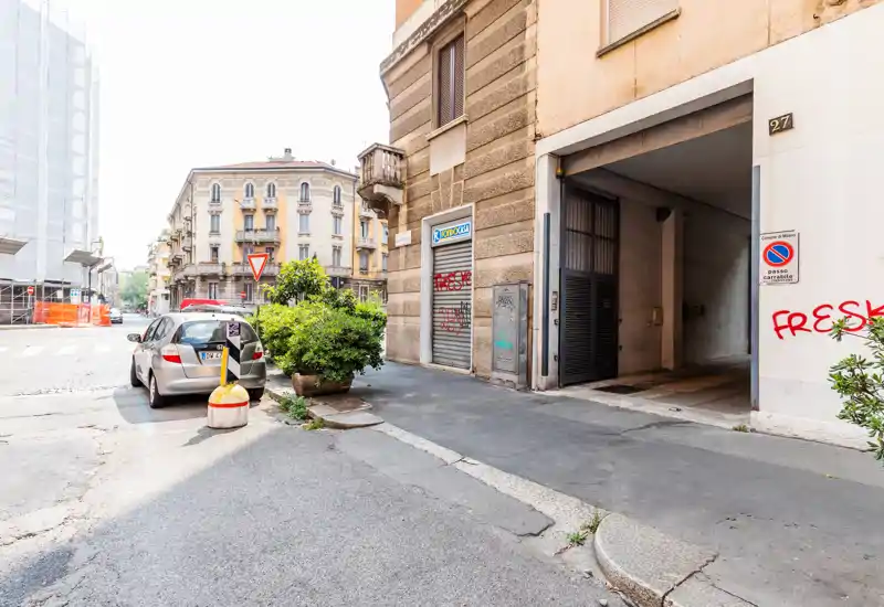 Street-level exterior showing a building entrance, garage/driveway opening and parked car on a sunny urban sidewalk.