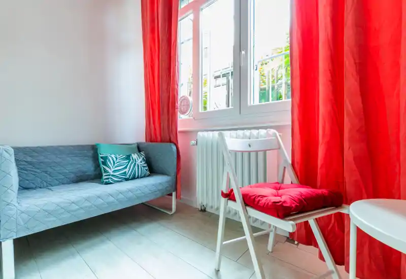 Small living area with a compact sofa, folding chair and vibrant red curtains that let in bright natural light — a cozy, well-lit sitting corner ideal for short stays.