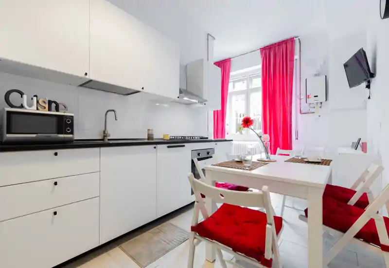 Bright modern kitchenette with white cabinetry, black countertop and a small dining table with red-cushioned chairs; vivid red curtains and decorative accents create a fresh, welcoming space.