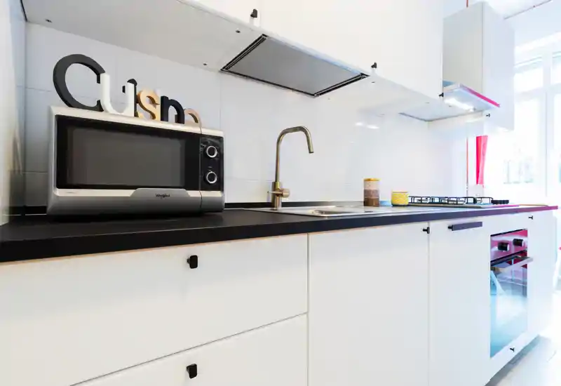 Bright modern kitchen with white cabinetry, black countertop, built-in oven, microwave and a sink beneath contemporary faucet. Clean, well-lit space ideal for rental listings showcasing a compact, stylish cooking area.