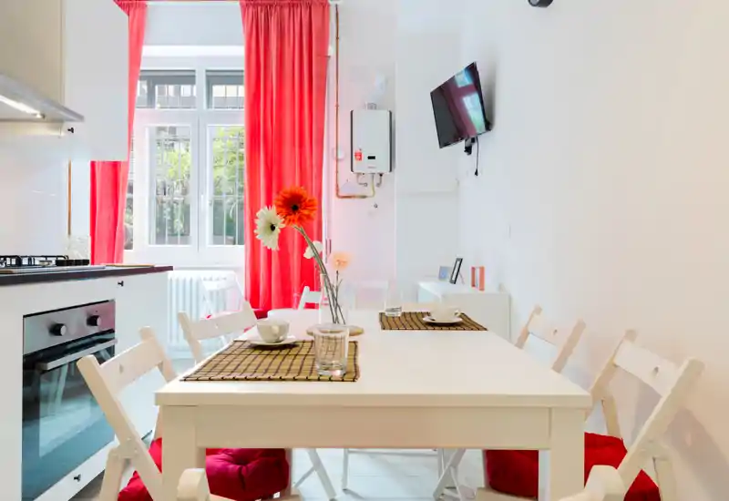 Bright, well-staged dining area connected to the kitchen: white table and chairs, red cushions and curtains, decorative flowers, and table settings for two. Ideal for listing photos highlighting a cozy eating space.