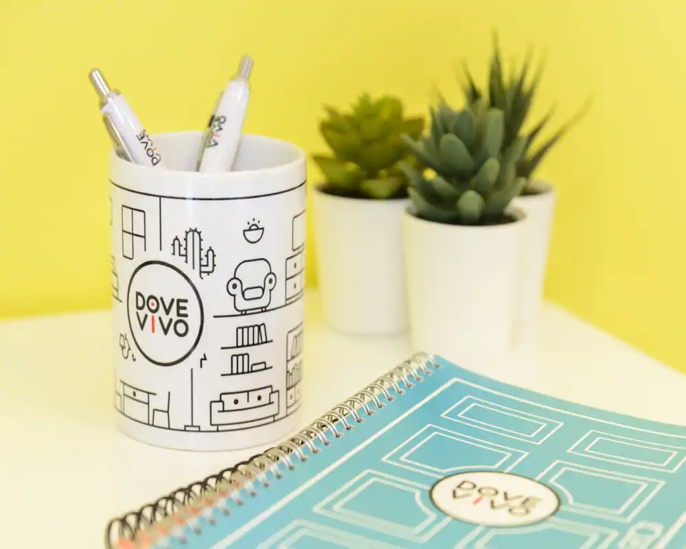 Primo piano di oggetti da scrivania: porta penne brandizzato, quaderno a spirale e piccoli cactus in vaso su uno sfondo giallo. L'immagine mette in risalto accessori per la scrivania e il brand della cancelleria.
