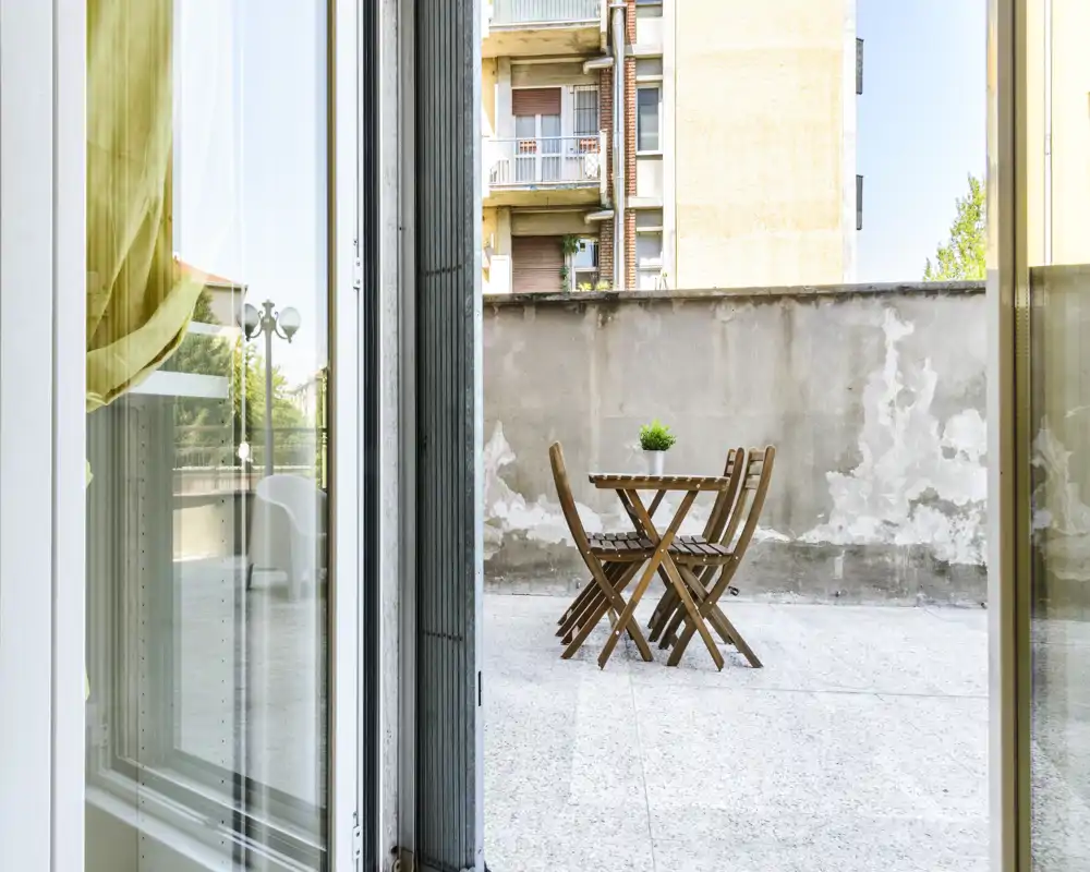 Terrazza privata soleggiata con un piccolo tavolo in legno e sedie pieghevoli, perfetta per il caffè mattutino. Pavimento piastrellato e un muretto basso offrono un angolo esterno tranquillo in città.