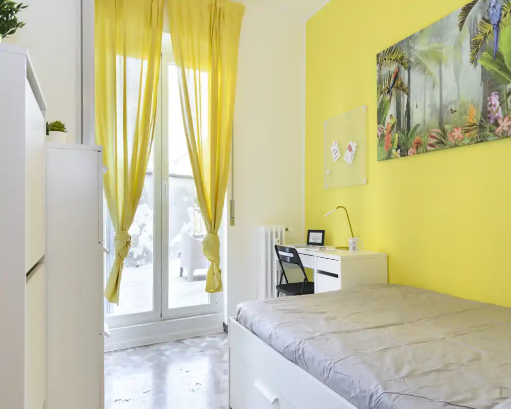Camera da letto luminosa con letto matrimoniale, mobili bianchi e una scrivania vicino a una grande finestra con tende gialle — ariosa, ben allestita e ideale per annunci.