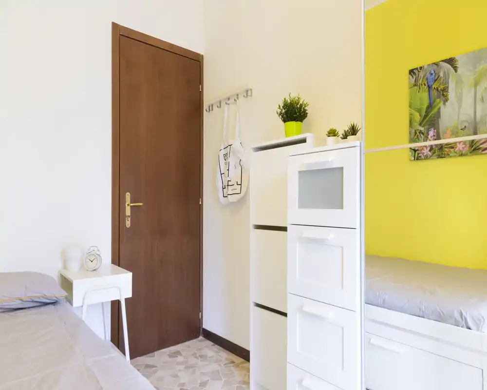 Camera da letto luminosa e ordinata con letto ben visibile, comodino e piante decorative; la parete gialla dona personalità e un’atmosfera accogliente.