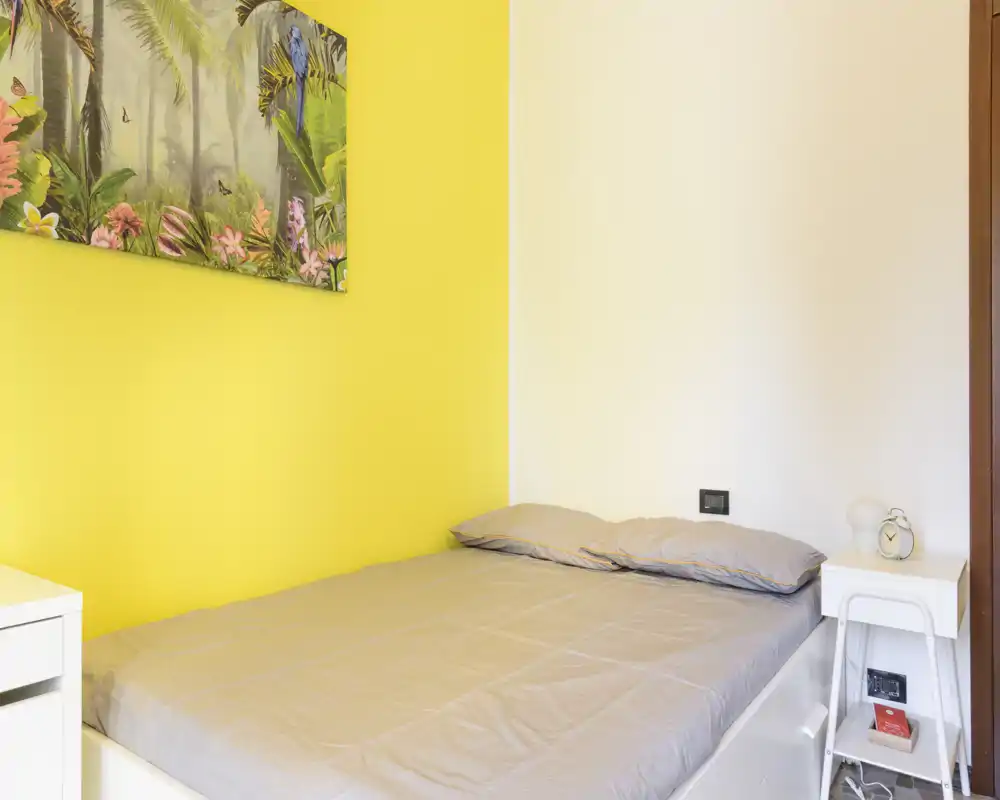 Camera da letto luminosa e compatta con un letto singolo ben rifatto contro una parete accentata gialla e un comodino con sveglia — ideale per un soggiorno accogliente per una persona.