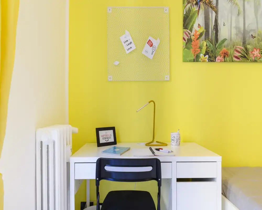 Angolo di camera da letto luminoso con scrivania bianca, sedia e parte del letto visibile a destra; pareti gialle vivaci, lampada da scrivania e decorazioni rendono l'area studio ordinata.