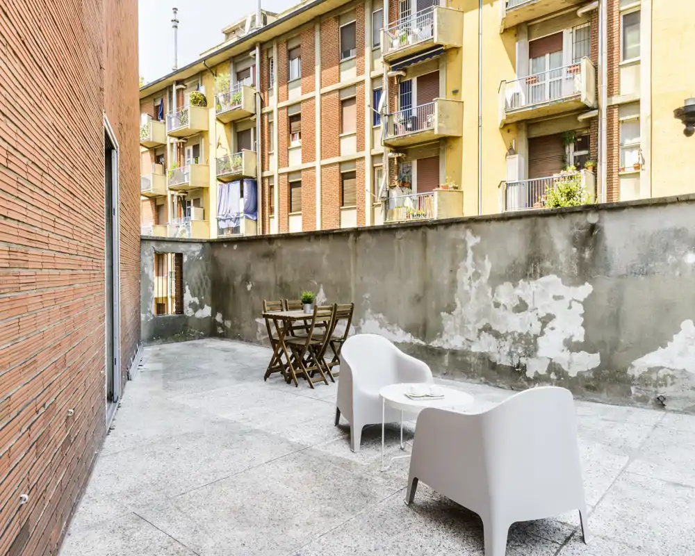 Terrazza privata soleggiata con posti a sedere per quattro: due poltrone moderne e un tavolino in primo piano e un set da pranzo in legno sullo sfondo. Spazio pavimentato e delimitato da un muro alto, ideale per pasti all'aperto o un caffè in città.