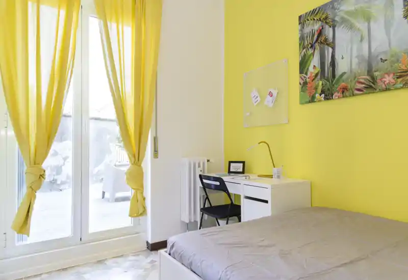 Bright, modern bedroom with a comfortable bed, a small desk and large glass doors that open to the outside; cheerful yellow accents and artwork create a sunny, inviting atmosphere.