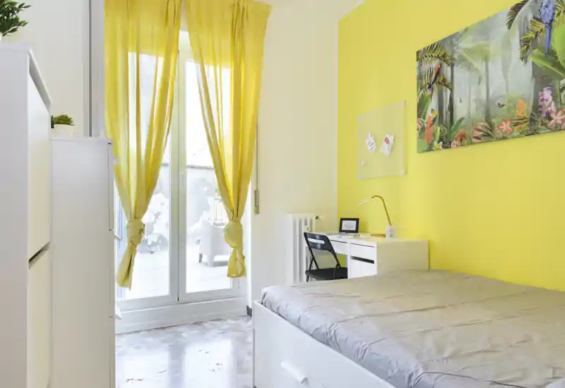 Bright bedroom with a double bed, white furniture and a study desk beside a large window with yellow curtains — fresh, airy and well staged for rental photos.
