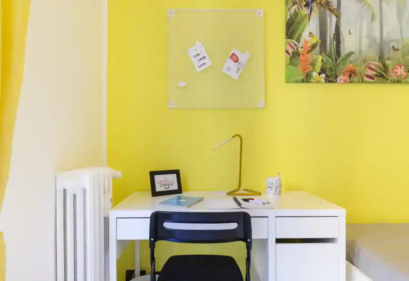 Small bright bedroom corner with a white desk, chair and a partial bed visible at the right; cheerful yellow walls, desk lamp and wall decor create a tidy study corner inside the sleeping area.