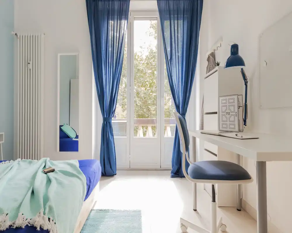 Camera da letto luminosa e moderna con un singolo letto ben rifatto, tessuti morbidi e una scrivania vicino alla porta-balcone illuminata dal sole con tende blu.