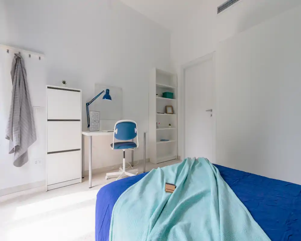 Camera da letto luminosa e moderna con letto visibile con tessili blu e coperta turchese; pareti bianche, scrivania compatta e scaffale per un aspetto pulito e minimalista.