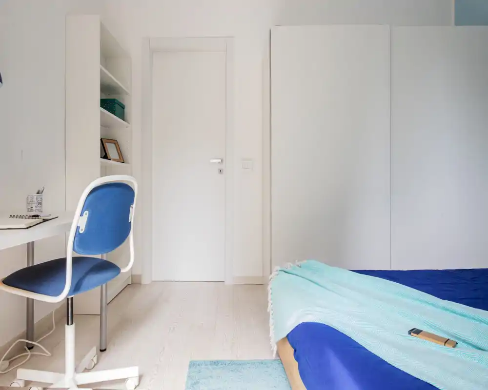 Camera da letto moderna e luminosa con letto rifinito in lino blu, un plaid turchese chiaro e una scrivania essenziale con sedia blu; arredo minimale e arioso.