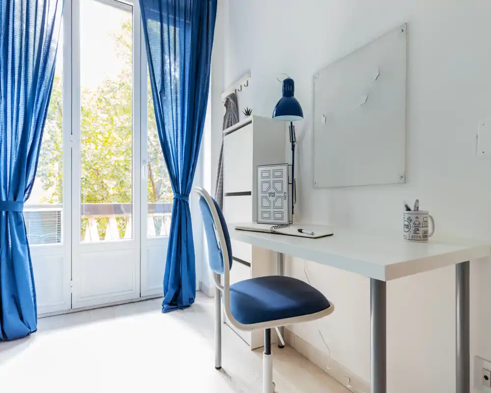 Angolo soggiorno/ufficio luminoso e ordinato con scrivania bianca e sedia e tende blu; ampie finestre a tutta altezza illuminano l'ambiente rendendolo ideale come postazione di lavoro.