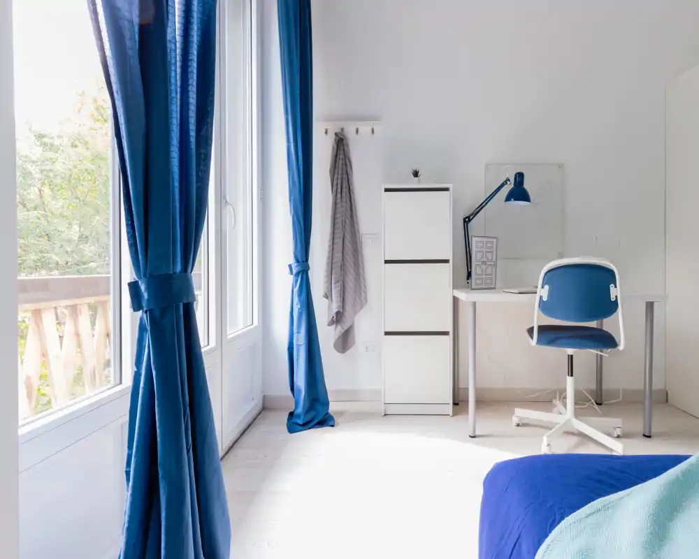 Camera da letto luminosa e minimal con ampie finestre, tende blu e copriletto blu; una scrivania compatta e una sedia creano un angolo studio funzionale.