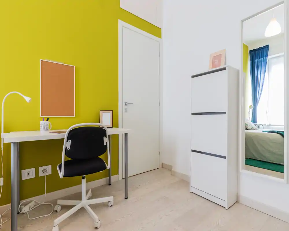 Rincón de dormitorio luminoso y moderno con un escritorio frente a una pared verde lima y un espejo que refleja parte de la cama y la ventana.