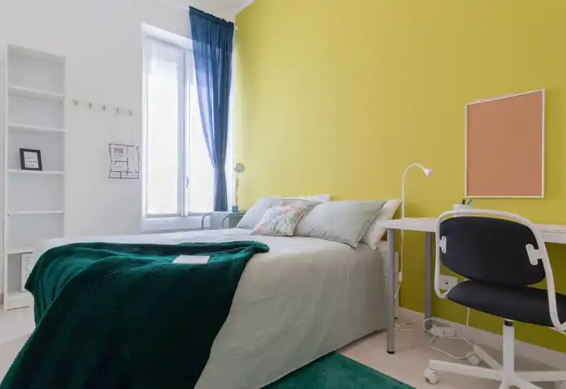 Dormitorio luminoso y moderno con cama doble, manta verde azulado y cojines decorativos, mucha luz natural desde la ventana alta y un pequeño escritorio.