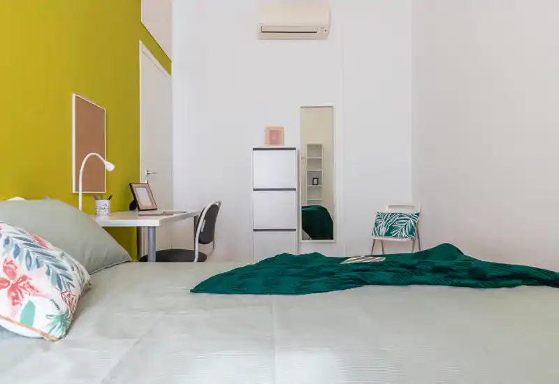 Dormitorio moderno y luminoso con cama bien hecha, cojines decorativos y manta verde; incluye escritorio y silla junto a una pared de acento llamativa.