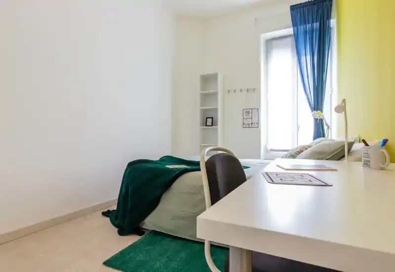 Dormitorio luminoso y ordenado con cama individual, escritorio y luz natural desde la ventana; textiles verdes y estantería sencilla para un ambiente funcional y tranquilo.