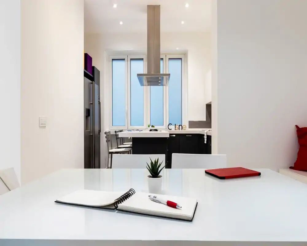 Cocina moderna y luminosa en concepto abierto vista desde la mesa de comedor/trabajo. Superficies blancas limpias, electrodomésticos de acero inoxidable y una pequeña planta decorativa crean un espacio funcional y fresco.