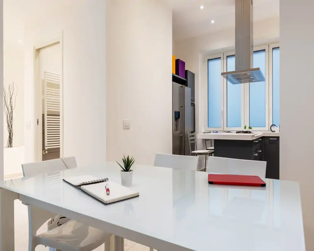 Zona de comedor moderna y luminosa con mesa blanca brillante, sillas y cocina abierta cercana; estilo minimalista y limpio, ideal para anuncios.
