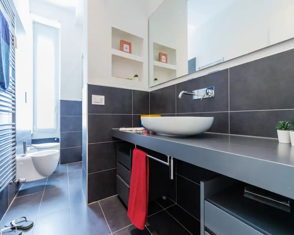 Baño moderno y bien iluminado con lavabo sobre encimera, grifo empotrado en la pared, bidé junto a una ventana alta y azulejos oscuros en paredes y suelo.