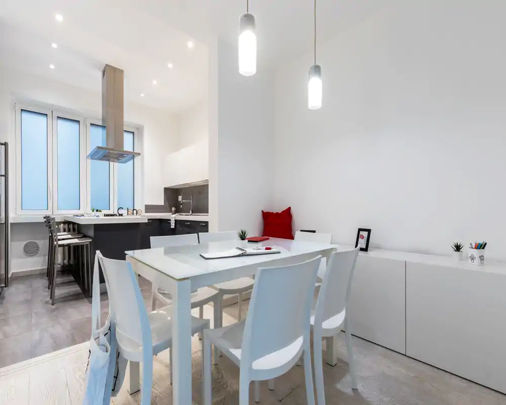 Zona de comedor moderna y luminosa con mesa de cristal, seis sillas blancas y lámparas colgantes; espacio abierto hacia una cocina compacta y almacenamiento minimalista.