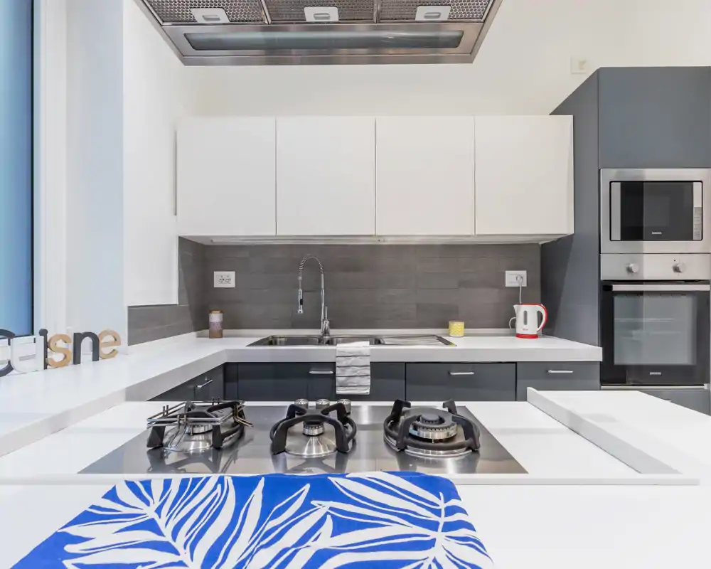 Cocina moderna totalmente equipada con placa de gas de acero inoxidable en la isla, encimeras blancas y muebles altos, horno y microondas empotrados y un grifo elegante. Luminoso, limpio y de diseño contemporáneo, ideal para anuncios.