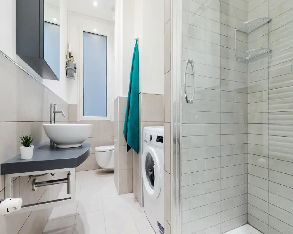 Baño moderno y compacto con ducha de cristal, inodoro suspendido, lavabo sobre encimera flotante y lavadora de carga frontal; azulejos claros y una toalla turquesa que aportan frescura.