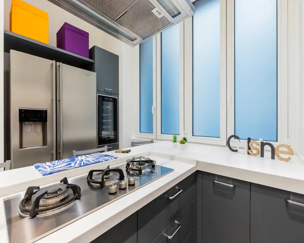 Cocina moderna y luminosa con encimera blanca, placa de gas integrada y frigorífico de acero inoxidable. Pequeñas plantas y cajas de colores añaden un toque contemporáneo.