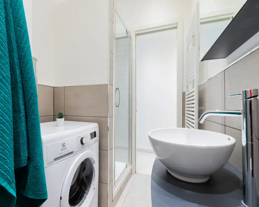 Baño moderno con lavabo tipo cuenco, grifo cromado, ducha con mampara de vidrio y lavadora; espacio luminoso, limpio y compacto.