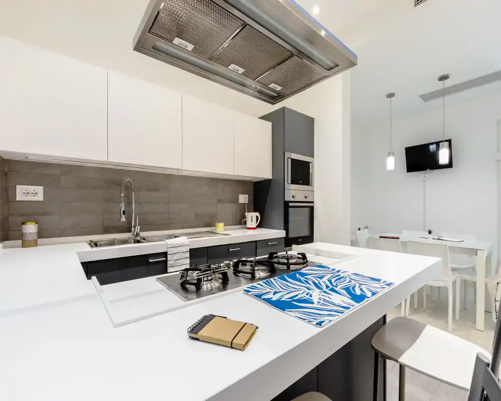 Cocina moderna y luminosa con isla blanca, placa de gas, campana extractora en techo y electrodomésticos integrados; se aprecia una pequeña zona de comedor al fondo.