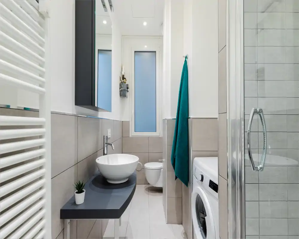 Baño moderno y compacto con lavabo sobre repisa flotante oscura, inodoro suspendido, ducha con puerta de vidrio, radiador toallero y lavadora — luminoso y bien acabado.