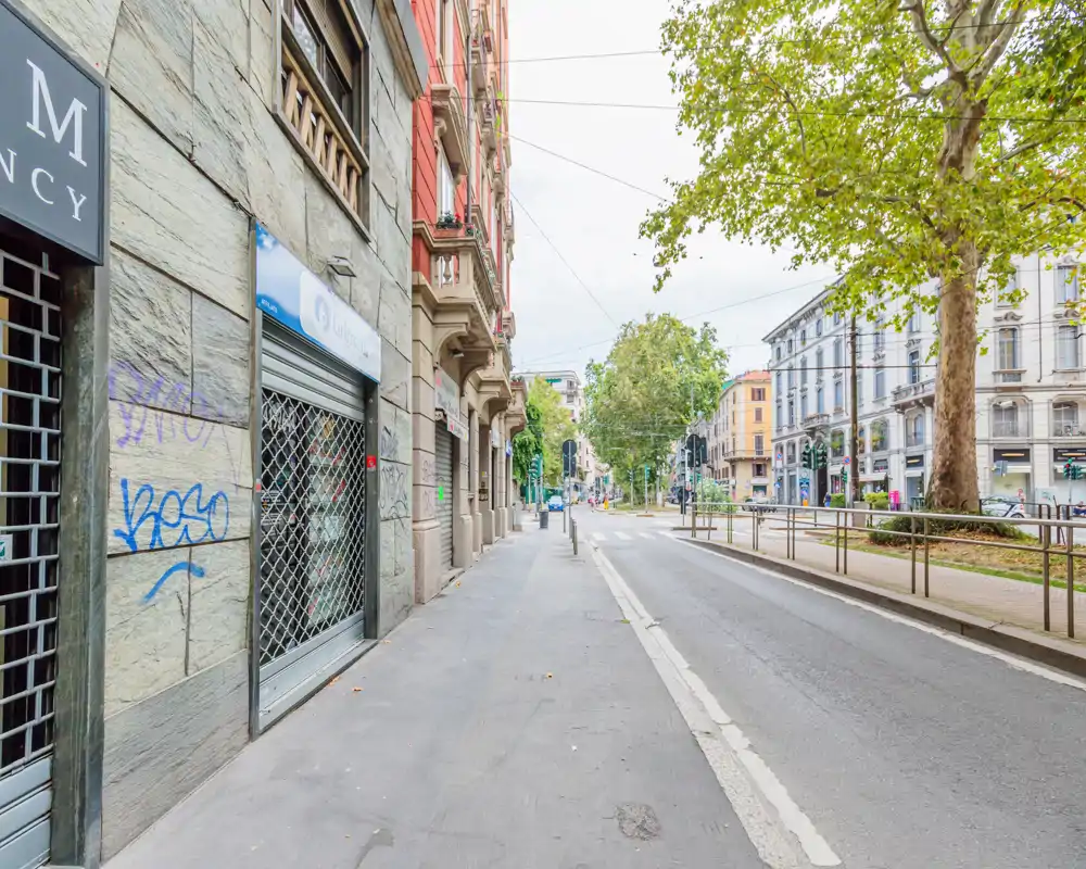 Vista urbana luminosa de la calle y la acera con comercios, árboles y líneas de tranvía — idónea para resaltar la ubicación y los servicios cercanos.