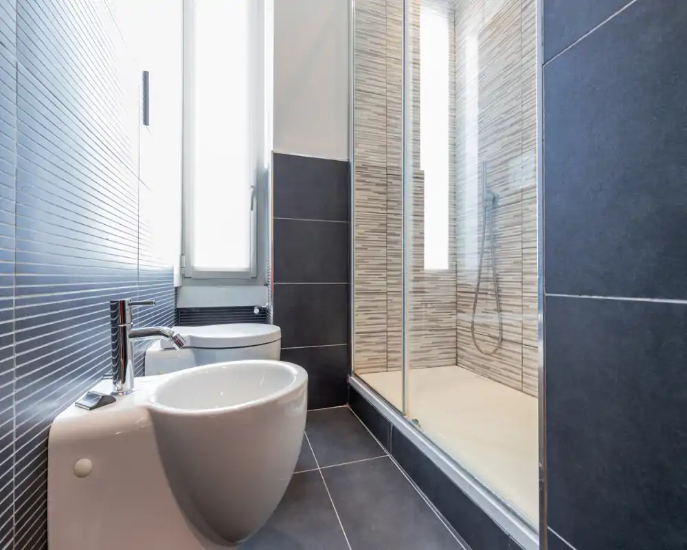 Baño moderno y luminoso con cabina de ducha, bidé e inodoro, suelos de baldosa oscura y decoración de mosaico vertical en la ducha.