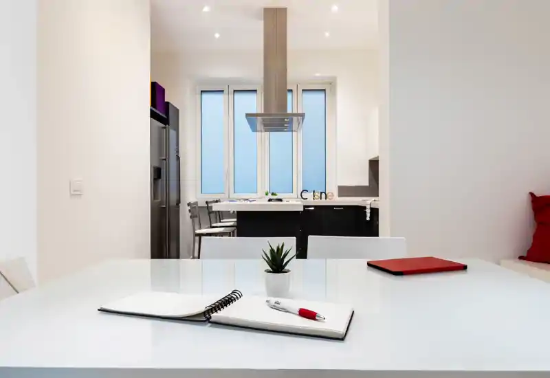 Cucina moderna e luminosa in open space vista dal tavolo da pranzo/lavoro. Superfici bianche pulite, elettrodomestici in acciaio e una piccola pianta decorativa rendono l'ambiente funzionale e fresco.
