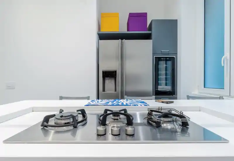 Cucina moderna e luminosa con piano cottura a gas in acciaio inox in primo piano e frigorifero a doppia porta con cantinetta sullo sfondo. Stile pulito e minimal con tocchi di colore dalle scatole.