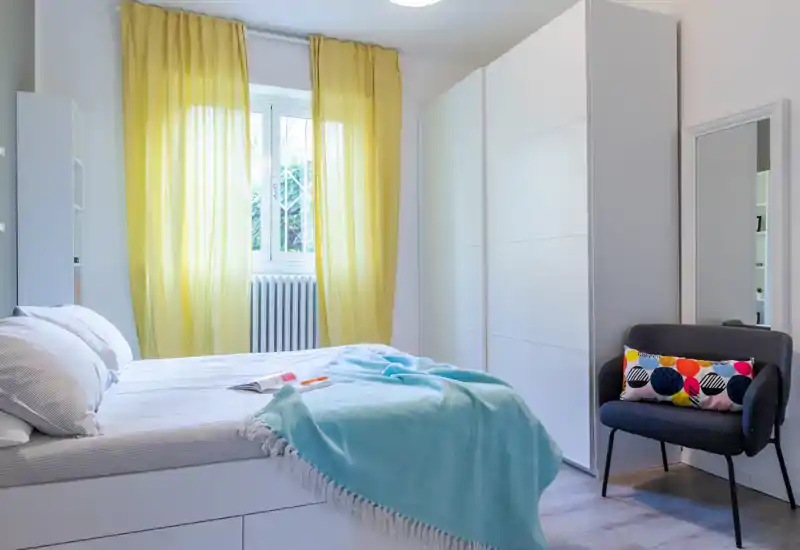 Bright, well-staged bedroom with a large bed, soft turquoise throw and sunny yellow curtains; a roomy white wardrobe and an accent armchair complete the space.