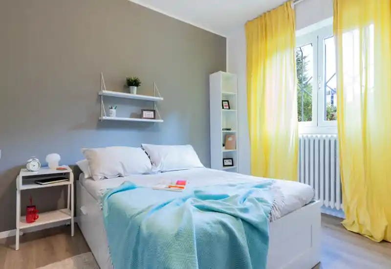 Bright, modern bedroom with a large white bed, soft blue throw and sunny yellow curtains; tidy bedside table and shelving create a fresh, welcoming atmosphere.