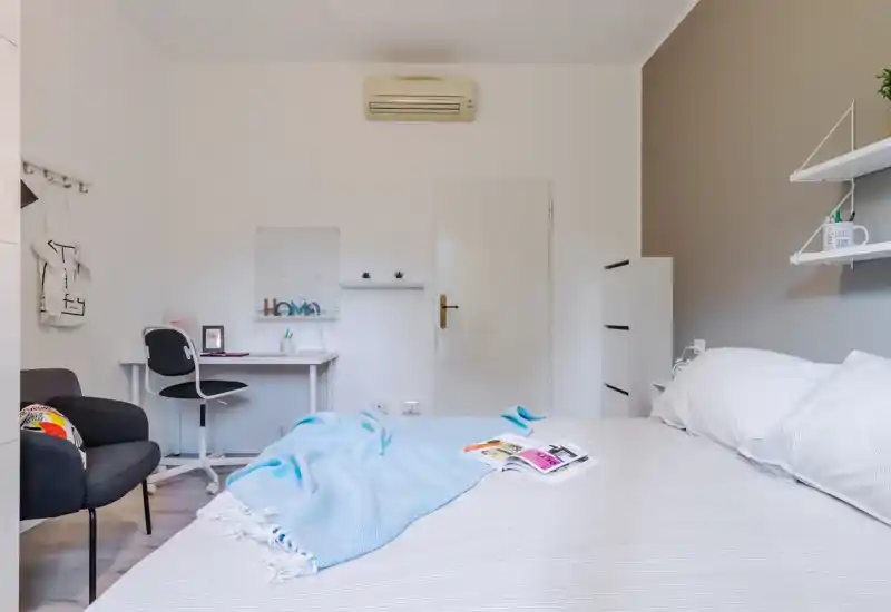 Bright, well-styled bedroom with a large bed in the foreground, soft linens and a cozy throw. A small desk and chair sit against the wall, making the room suitable for rest and light work.