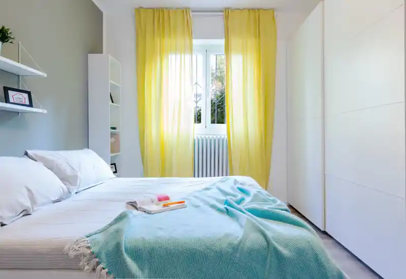 Bright, cozy bedroom with a large bed dressed in white linens and a turquoise throw, yellow curtains framing the window and a clean modern wardrobe.
