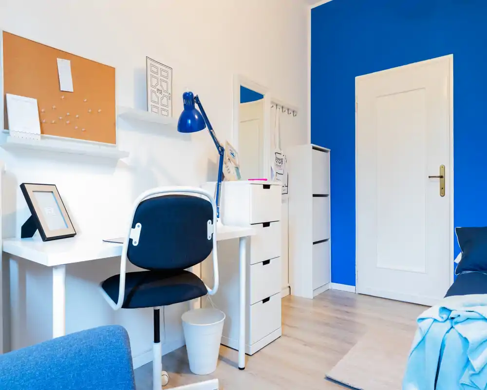 Bright, tidy bedroom corner with a white desk and blue-accented decor; a small portion of a bed is visible at the right edge, making this a functional and stylish sleeping/study area.