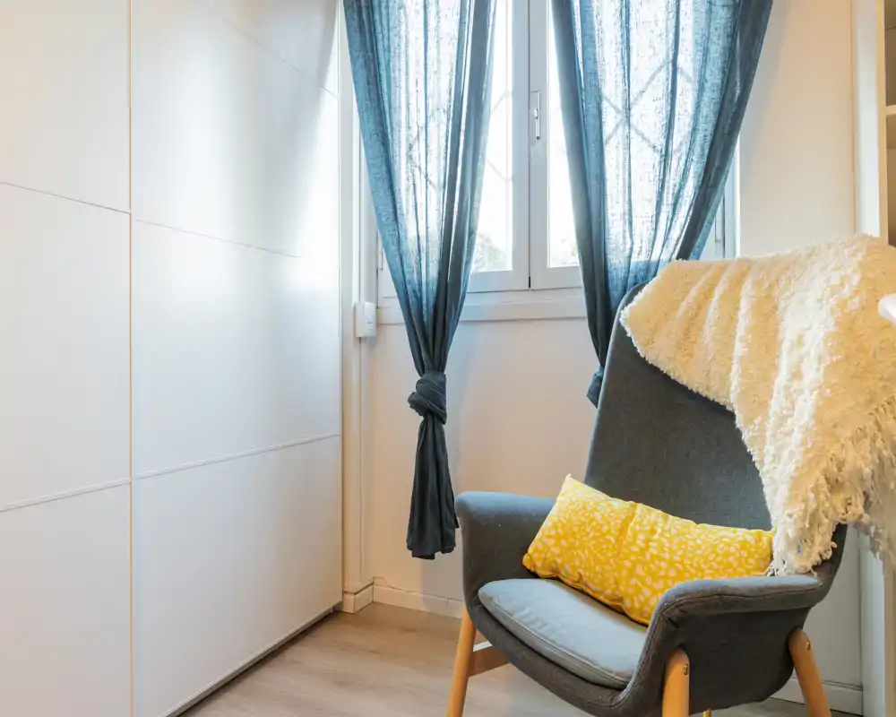 A bright, cozy reading corner with a modern upholstered armchair, soft throw and decorative yellow cushion beside a curtained window.