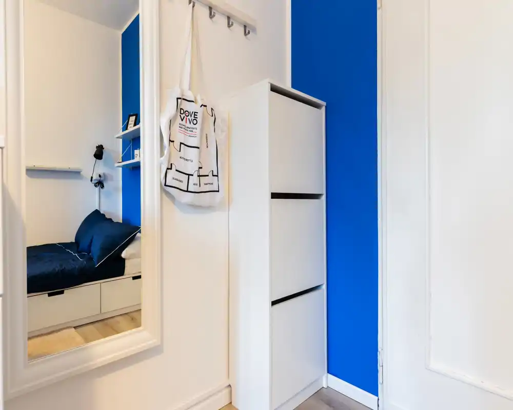 Compact bedroom view seen through a hallway mirror, showing a single bed with dark bedding, built-in storage drawers and a modern white shoe cabinet against a vivid blue accent wall.
