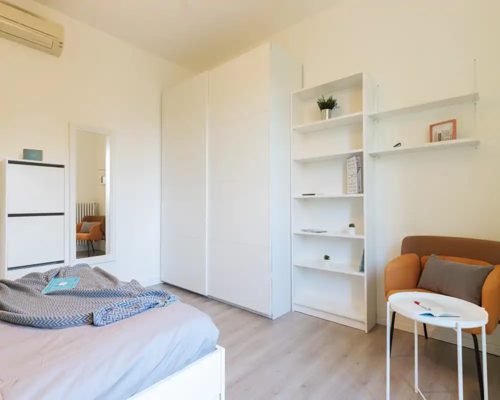 Dormitorio moderno y luminoso con cama visible, gran armario blanco, estanterías y un sillón acogedor — minimalista, aireado y bien iluminado.