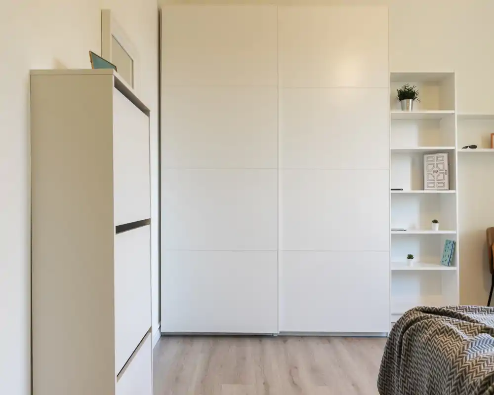 Rincón de dormitorio minimalista con un gran armario blanco y estanterías; una manta estampada en la esquina inferior derecha insinúa la cama fuera de plano. Líneas limpias y tonos neutros que transmiten una atmósfera moderna y serena.