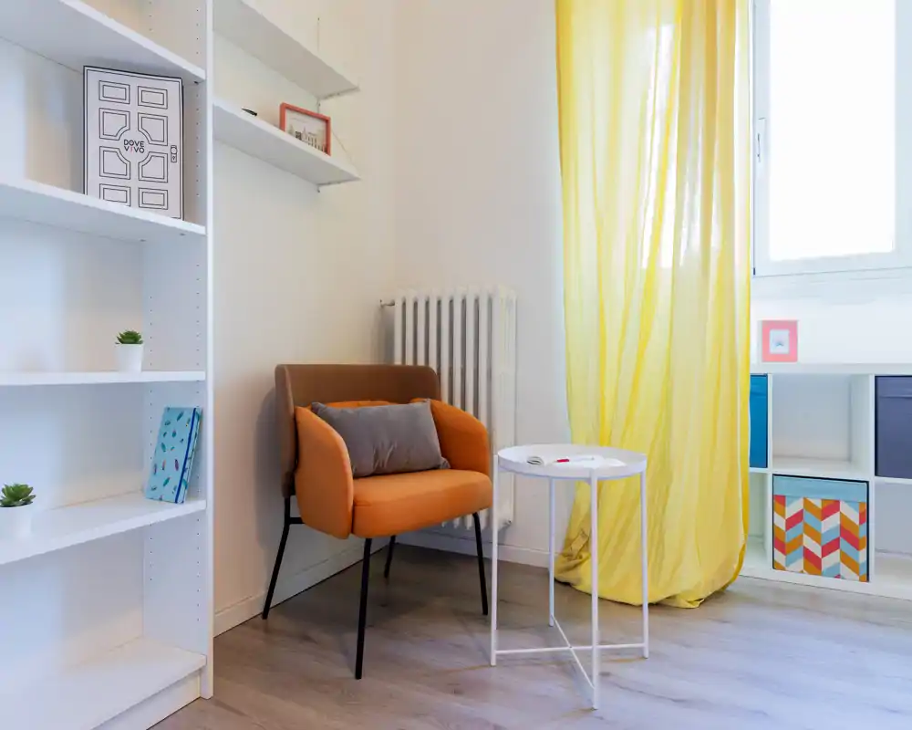 Rincón de salón luminoso y moderno con un cómodo sillón naranja, mesita blanca y estanterías — sensación ligera con luz natural a través de la cortina amarilla.