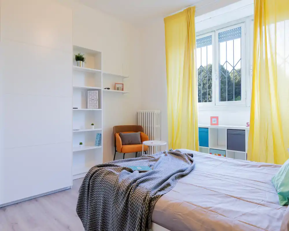 Dormitorio luminoso y acogedor con la cama en primer plano, textiles suaves y cortinas amarillas que dejan entrar abundante luz natural.