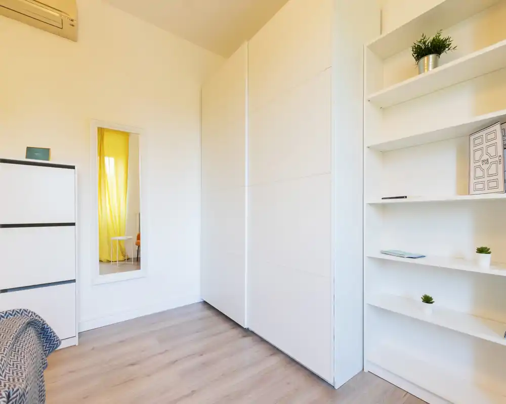 La imagen muestra una esquina de interior minimalista con armarios empotrados blancos, estanterías, una cómoda, un espejo en la pared y parte del suelo; no se aprecia una cama ni elementos claros de sala/cocina/baño para identificar el tipo de habitación.